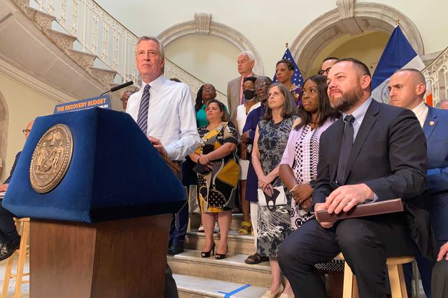 Mayor de Blasio at a lectern, with City Council Speaker Johnson seated next to him and other electeds behind them in the rotunda of City Hall